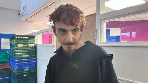 Kate Bradbrook/ BBC A person stands inside a brightly lit room at the Hope Centre’s day facility, wearing a dark coat. Behind them, tall stacks of colourful plastic crates are arranged against the wall, alongside white partition panels and windows looking into adjoining rooms. Fluorescent ceiling lights and vibrant interior colours create a busy, functional setting typical of a community support space.