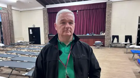 Kate Bradbrook/BBC Sean is pictured standing inside a large church hall that has been converted into a winter night shelter. Rows of grey fold‑out camp beds, each topped with a pillow and neatly arranged across the wooden floor, fill the room. Behind them, a stage with deep red curtains runs along one side of the hall, and personal belongings can be seen placed on benches around the edge of the space. The high ceiling, exposed beams and brick walls give the room a community‑hall feel, while the lighting above illuminates the scale of the operation set up to support people sleeping rough.