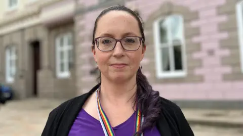 Deputy Louise Doublet in a purple top and glasses stands in front of a pink granite building, Jersey's States Assembly.