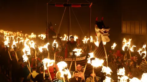 Reuters Jarl Squad Vikings take part in a procession. Some are riding in the long boat. All those taking part are carrying flaming torches.

