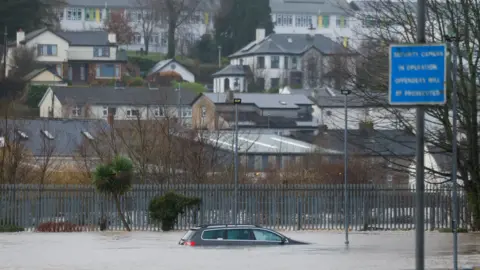 Reuters A wide shot shows a grey estate car submerged up to the windows in floodwater - in the distance a grey metal fence can be seen and behind that a range of different buildings including homes and offices. 