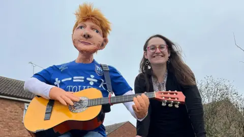Sharon Kulesa A woman - Sharon Kulesa - smiling at the camera while standing next to an effigy of Ed Sheeran which is wearing an Ipswich Town top and holding a guitar.