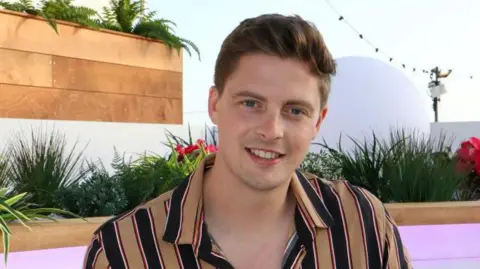 Getty Images Dr Alex George on the set of Love Island. It is a head and shoulders shot and he has a beige and black striped shirt on. 