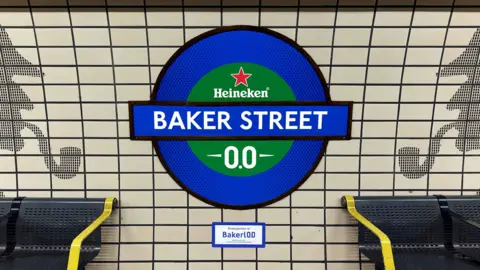 Heineken A London underground a wall sign reading “Baker Stret”, with  Heineken branding inserted. The TFL sign is blue anbd green . Bakerloo is spelled with "0.0" instead of "oo" at the end