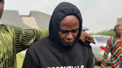 BBC Adeniyi Kayode wears a black hoodie an looks downwards. Another man, out of shot, puts his arm round Mr Kayode