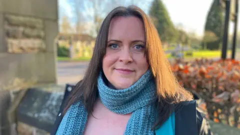 BBC Jen Dinelow, who paid higher "non-resident" charges for her parents' burials, stands outside a cemetery. She has long, light brown hair and is wearing a light blue scarf over a pink top.