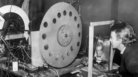 Getty Images A black and white image of John Logie Baird, the inventor of television, with his transmitting station, transmitting disc.