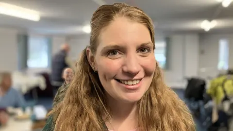 BBC Vikki has long light brown hair which is tied back but also flows over her shoulders. She has brown eyes. She is smiling for the camera.