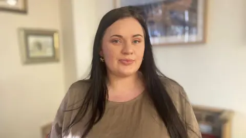 BBC Fiona Goddard, who has long dark hair and wears a brown top, looks straight at the camera.