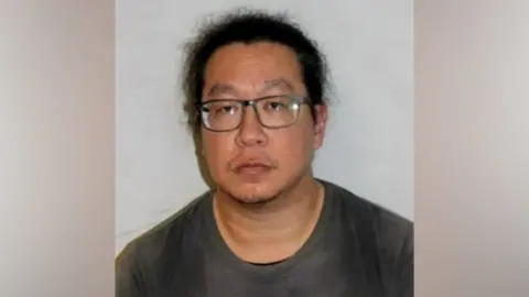 Metropolitan Police Vincent Chan police custody image. He wears glasses and a dark grey top and looks into the distance. 