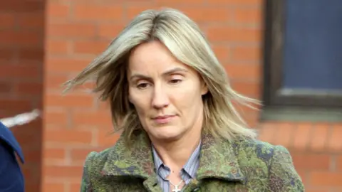 PA Media Hazel Stewart, with shoulder length blonde hair, wears a green floral coat with a blue and grey shirt. Behind her is a red-brick building and dark translucent window.