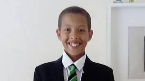 Pacemaker Press Noah smiling at the camera as he stands against a white wall. He is wearing his school uniform which is a black blazer, green and black striped tie and a white shirt. He has short black hair.