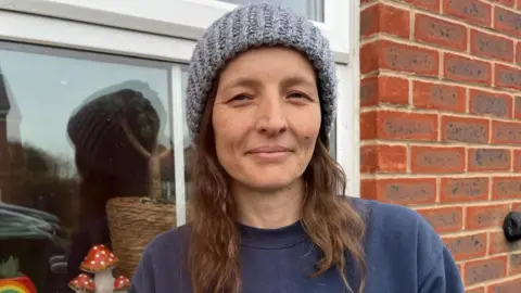 BBC Lucy in a grey hat. She has brown hair and is also wearing a blue jumper. She is stood in front of a window.