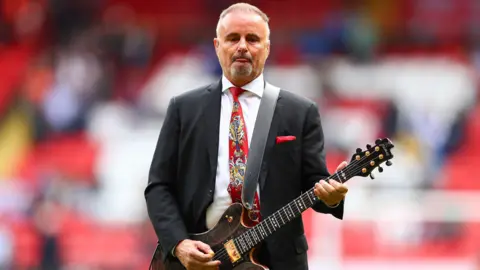 Getty Images A bearded man wearing a red tie and suit inside a football stadium. He is playing a guitar