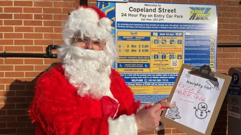 BBC Father Christmas holding a naughty list that says "Excel Parking"