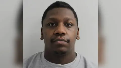 Met Police A mugshot style image of a man. He has a blank expression and is staring down the camera. He is wearing a plain grey jumper and has a moustache and short hair