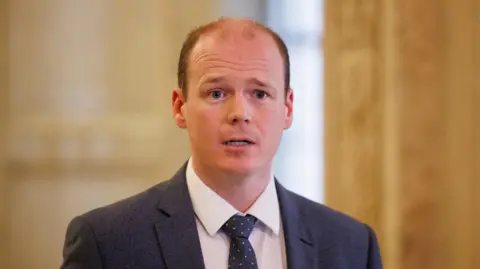 PA Media A man with short red/brown hair looks to the right of the camera. He wears a dark grey suit, white shirt and Navy tie. Behind him is an out of focus dark cream wall. 