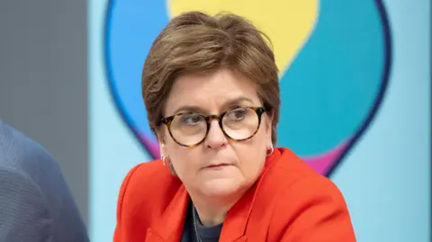 PA Media Woman wearing glasses and a red blazer, seated at a table and looking to her left, with a blurred colourful background behind her.