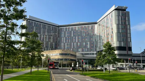 A modern hospital building with grey and coloured panels with a road and trees in the foreground