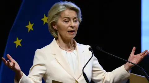 Getty Images European Commission President Ursula von der Leyen wearing cream suit stands in front of the European Union flag