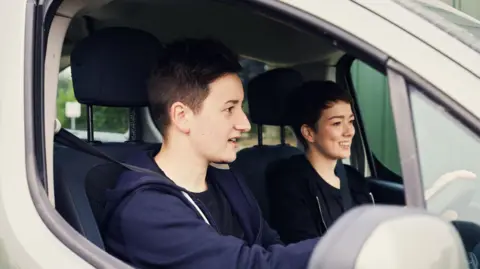 Getty Images A young learner driver. He has short, dark hair and is wearing a navy hoodie and top. In the passenger seat is a young woman with short dark hair, also wearing a dark hoodie and top.