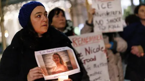 TOLGA AKMEN/EPA/Shutterstock A woman wearing a dark jacket with a hood over her head holds a candle and a picture of a Minneapolis woman fatally shot by an agent of ICE, the US  Immigration and Customs Enforcement agency