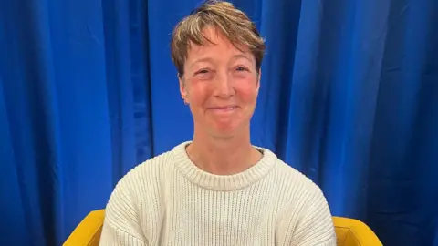BBC Nicky Perfect looks at the camera and smiles. She is wearing a cream wool jumper and sits on a yellow armchair. The backdrop is a blue curtain