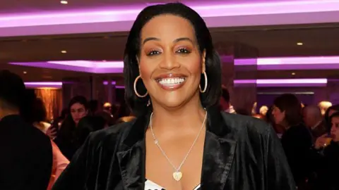 Getty Images A head and shoulders shot of Alison Hammond in a black jacket posing in a room lit with purple. She has her dark hair down but combed back from her fae and wears silver hooped earrings and a heart pendant necklace. Other women can be seen chatting in groups behind her.