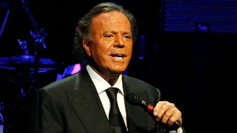 Getty Images Julio Iglesias performs 'Julio Iglesias In Concert At Gran Teatre del Liceu' In Barcelona on July 4, 2012 in Barcelona, Spain. He is wearing a black suit and tie and is holding a microphone.