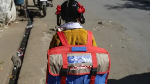 Getty Images A view from the back of a young girl, dressed in a school uniform and carrying a school bag, walking on a street in India.