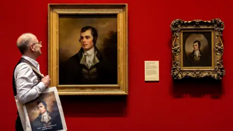 PA Media Dr Bill Zachs is looking at the lost portrait of Robert Burns, by the artist Sir Henry Raeburn, which is on display at the National Galleries Scotland next to the original Burns portrait by Alexander Nasmyth. Zachs is bald and wearing a red, yellow and dark checked waistcoat with a white shirt and mustard-coloured glasses. He is carrying a bag with the portrait on it, saying The Lost Portrait 