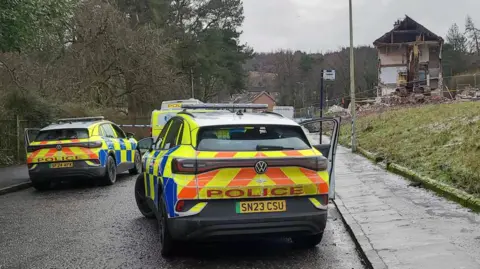 Watchtower Media Two police vehicles parked on a residential road near a severely damaged house, with debris visible and trees lining the street