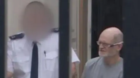 BBC Two men are in the picture. The one on the right is John Garrett O'Sullivan. HE is baldin with a grey beard and glasses. He is wearing a grey, round neck top. He is beside a man who has his face blurred and is wearing a white shirt and tie.