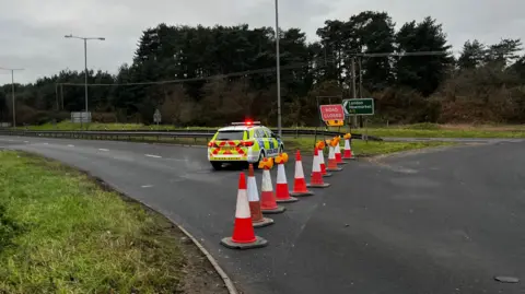 Jack Maclean/BBC A police car with flashing lights parked across a dual carriageway which has been blocked off to traffic by orange cones.
