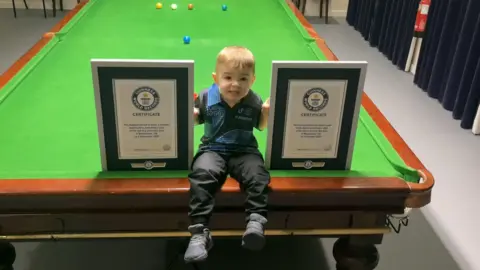 PA Media Jude, aged two, who has light brown hair and is wearing a blue t-shirt and black trousers, is sitting on the edge of a snooker table smiling next to two certificates.
