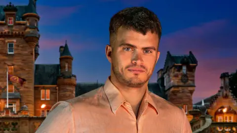 BBC/PA Traitors contestant Matthew Hyndman, with short brown hair and stubble, wearing a light pink shirt. He is standing in front of the Traitors castle and it is nighttime. 