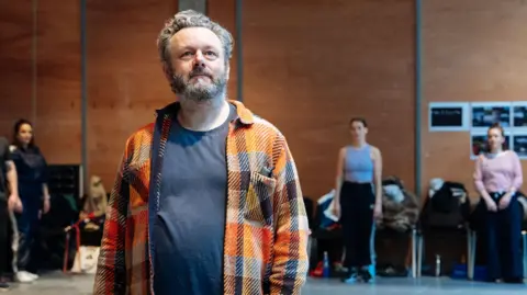 Helen Murray Michael Sheen in a checked shirt and T-shirt in the foreground, with other actors at the back of the rehearsal room
