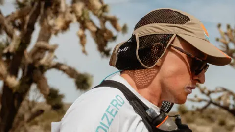 @adrianvarzaru Jonty Brown is wearing a brown cap and sunglasses and is wearing a white t shirt with a black sack around his neck containing water