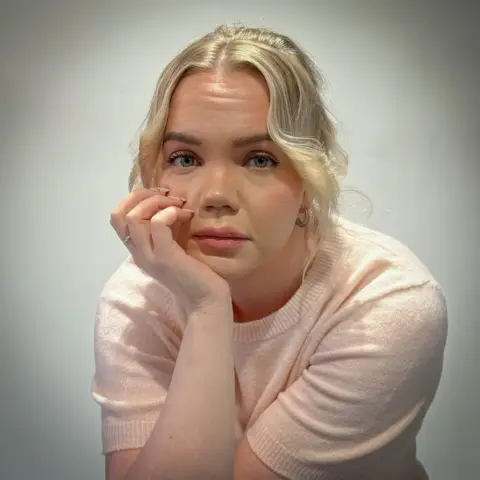 Elina Tossavainen Meri-Tuuli Auer looking towards the camera, she is seated with her head resting in her hand. She has blonde hair and is wearing a light pink short sleeved woollen top.