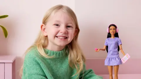 Mattel A young girl with blonde hair, smiling. She has a light green knitwear jumper on and is next to a Barbie doll which has ear defenders on and is holding a card and  a fidget spinner.