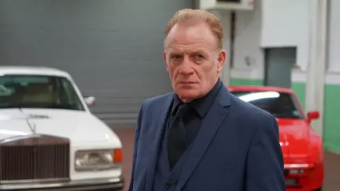 S4C Mark Lewis Jones, wearing a blue two piece suit with a dark shirt. He has light brown/ginger hair. Pictured behind him are two cars; one white and one red.