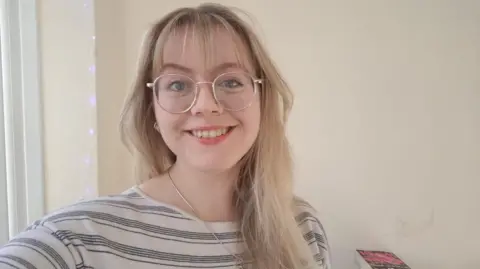Catherine Thoyts Catherine smiles at the camera, with long blonde hair, glasses, and a white top with black stripes. In the background are fairy lights and a pile of books.