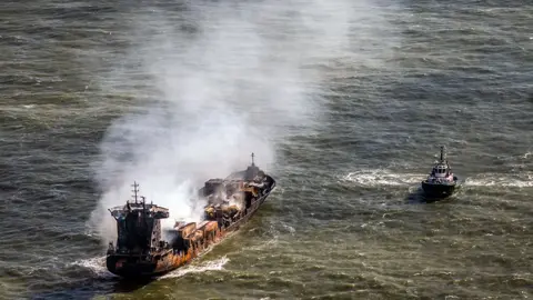 PA Media The Solong being shadowed by another ship. Smoke is coming out of the vessel and the hull and superstructure has burn marks across it 