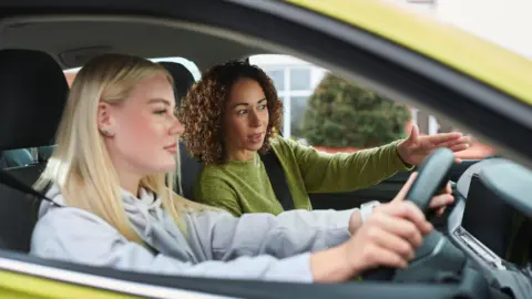 Getty Images A young girl with long blond hair is behind the wheel of a yellow car. There is a woman beside her, with curly hair and a green jumper, giving her instructions.