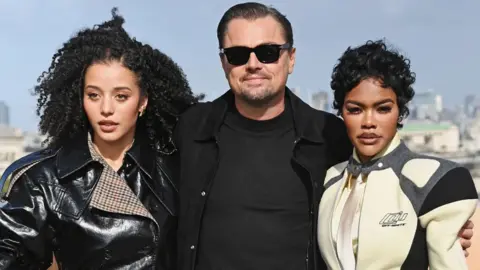 Getty Images Chase Infiniti, Leonardo DiCaprio and Teyana Taylor attend the London photocall for One Battle After Another in London in September. Infiniti is wearing a black shiny jacket, DiCaprio has a black jacket and T-shirt on and Taylor has on a racing-style cream, grey and black jacket.