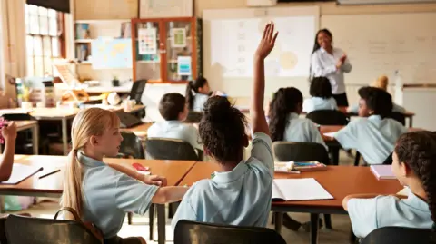 Getty Images The image shows the rear view of girl raising her right hand while sitting next to friend during a lecture in a full classroom.
