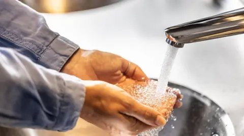 Getty Images Water running out of a tap onto a person's hands which are cupped. You can only see the grey sleeves of their shirt.