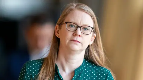 PA Media A woman wearing glasses and a green polka-dot top looks ahead with a neutral expression, standing indoors with a blurred background.