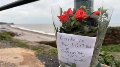 BBC A floral tribute tied to a pole. In the distance there is a beach. The flowers are red and there is a note in the flowers with the words 'Remembering those lost at sea on Christmas Day 2025. We will remember them.'