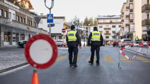 EPA/Shutterstock Two police officers wearing high visibility jackets walk on a cordoned off street near the site of the fire in the Crans-Montana ski resort.